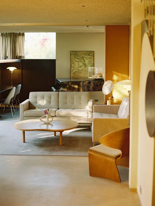 A modern living room interior featuring a neutral-toned sofa, wooden coffee table, and a plywood chair in a bright, naturally lit space.