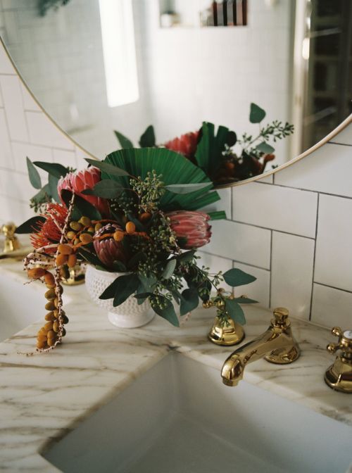 Elegant floral arrangement in a white vase with green leaves, featuring King protea flowers on a marble countertop in a bathroom.