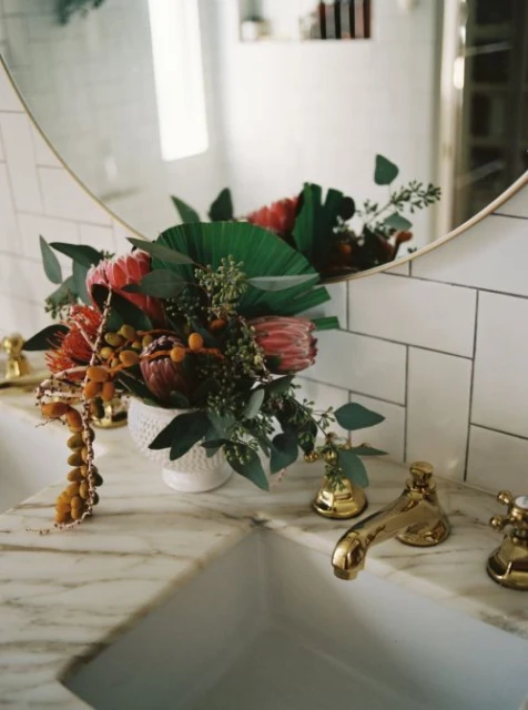 Elegant floral arrangement in a white vase with green leaves, featuring King protea flowers on a marble countertop in a bathroom.