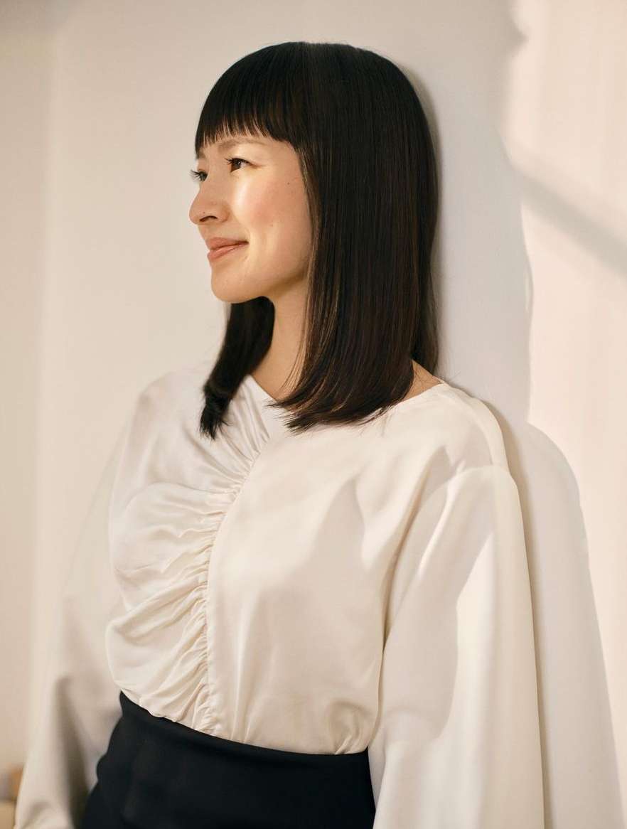Portrait of an adult female wearing a white long-sleeve blouse and black skirt, leaning against a white wall with soft, natural lighting.