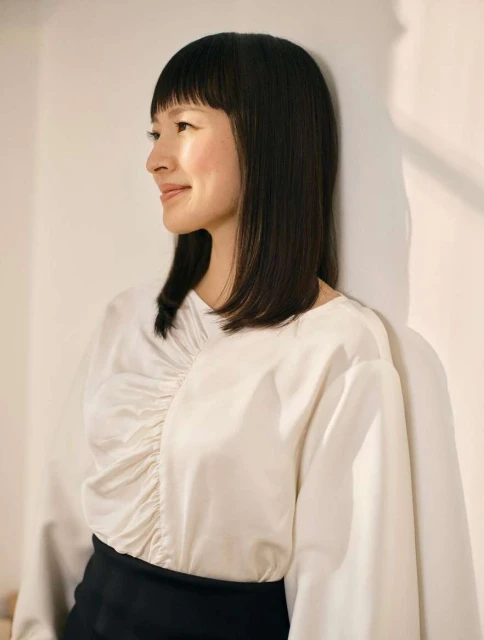 Portrait of an adult female wearing a white long-sleeve blouse and black skirt, leaning against a white wall with soft, natural lighting.