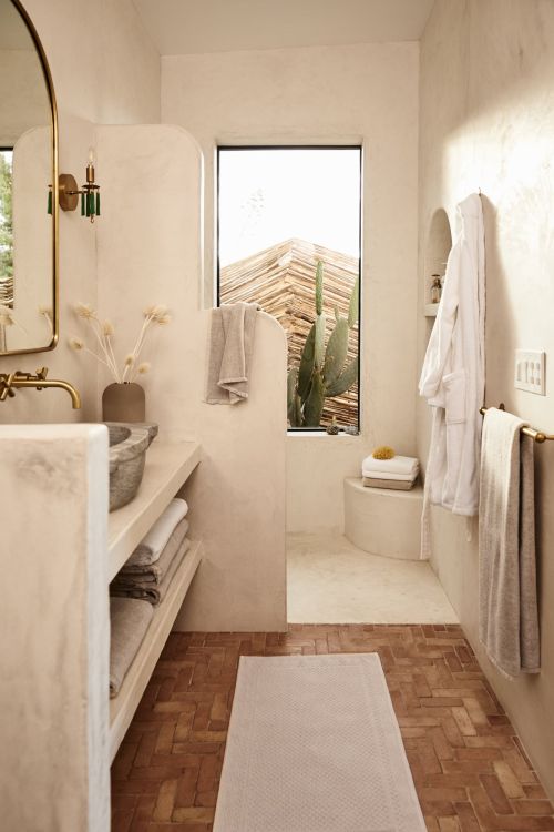 Neutral toned bathroom features stone sink, gold fixtures, stacked towels, white bathrobes, and cactus seen through window.