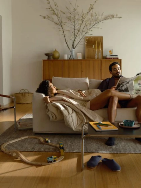 Couple relaxing on a modern couch in a bright living room with a newspaper, blanket, and wood toy train set on a rug.