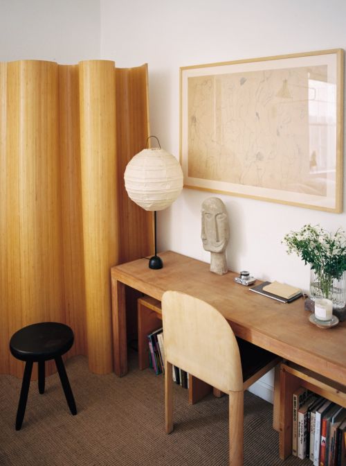 Home office interior with a light wood desk featuring a paper lantern style lamp, artwork, and a black stool next to a wood screen.