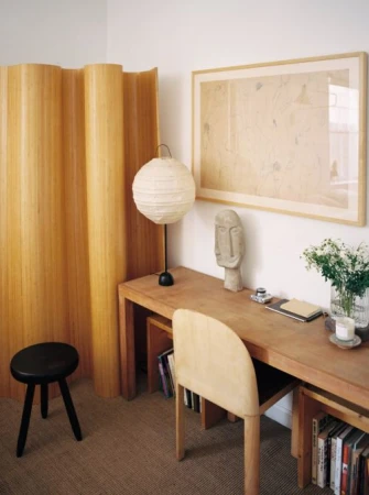 Home office interior with a light wood desk featuring a paper lantern style lamp, artwork, and a black stool next to a wood screen.