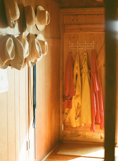 Hallway with wooden walls features straw hats, blue lanyards, rack with yellow and pink raincoats, creating a cozy, organized aesthetic.