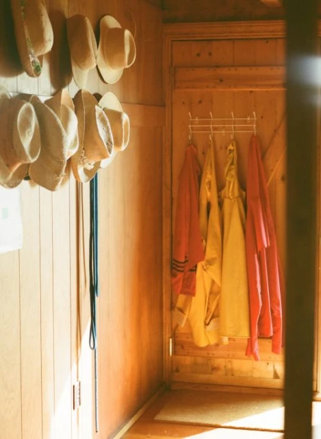 Hallway with wooden walls features straw hats, blue lanyards, rack with yellow and pink raincoats, creating a cozy, organized aesthetic.