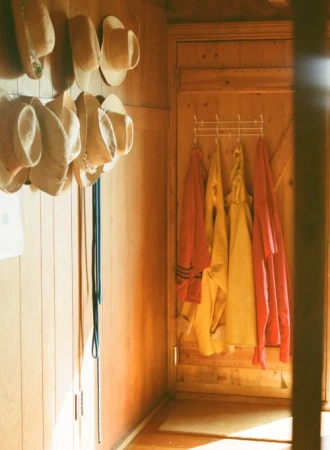 Hallway with wooden walls features straw hats, blue lanyards, rack with yellow and pink raincoats, creating a cozy, organized aesthetic.