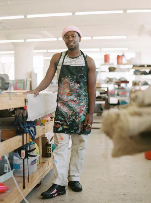 Smiling artist wearing paint-splattered apron, stands in art studio, light-filled space with art supplies, tools, and pink hat. 