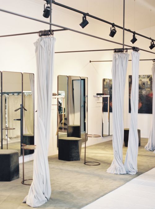 Interior view of a bright, modern clothing store featuring draped curtains, fitting rooms with mirrors, and spotlight track lighting.