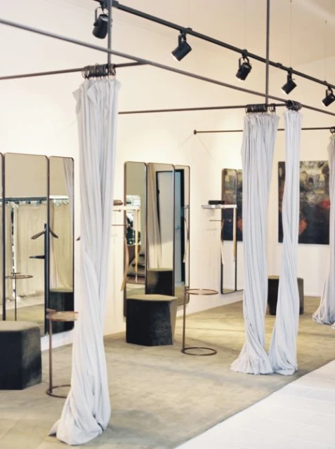 Interior view of a bright, modern clothing store featuring draped curtains, fitting rooms with mirrors, and spotlight track lighting.