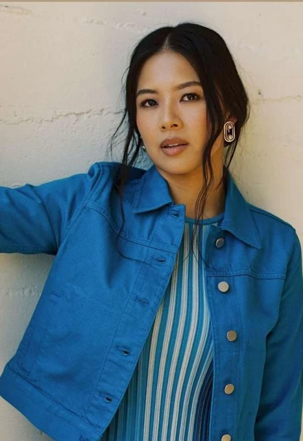 Attractive Asian woman in a blue denim jacket and striped top, looking towards the camera against a neutral-colored wall backdrop.