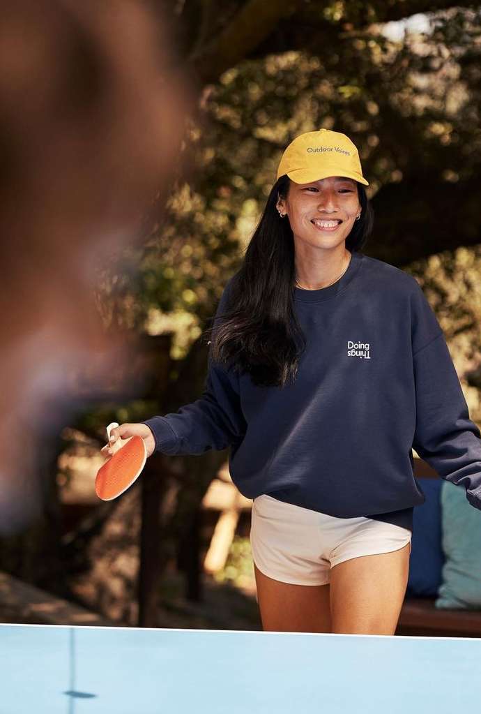 Smiling Asian woman in cap and sweatshirt playing ping pong outdoors with green foliage in the background during daytime.