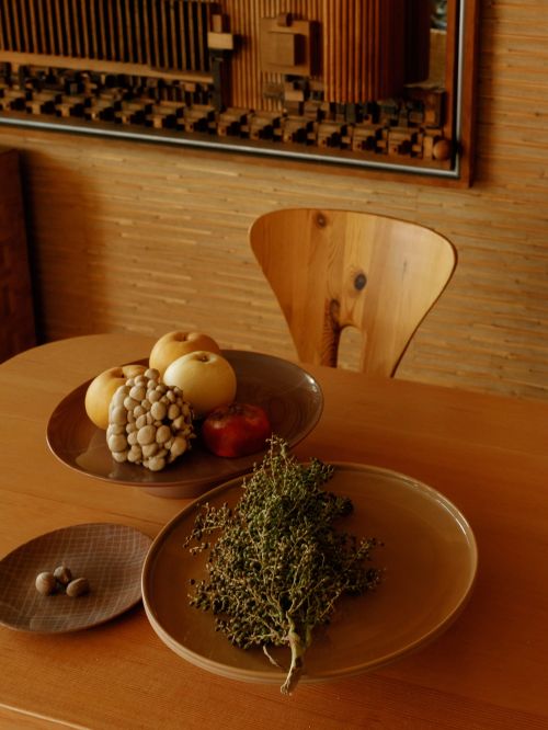 Still life composition with a wooden chair, fruit and decorative elements on a brown table against a light brown patterned wall.