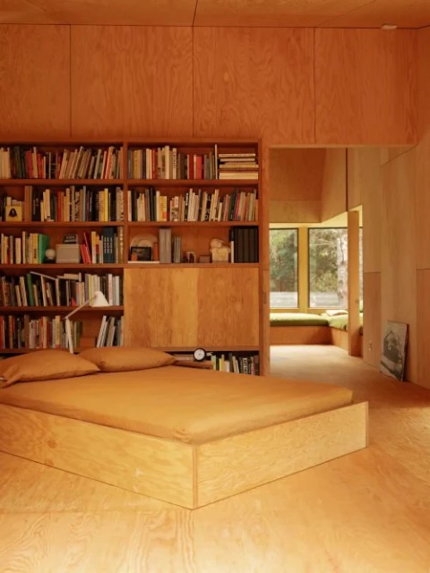 Plywood-clad bedroom with a platform bed and a built-in bookshelf featuring a small white desk lamp next to the bed pillows.