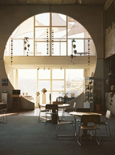 Sunlit interior featuring round tables and chairs, illuminated by natural light streaming through a large circular window.