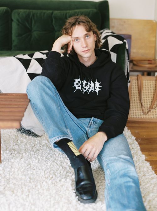 Adult male sitting on floor with black graphic hoodie, light-wash jeans, and dress shoes; white rug, green sofa, and woven bag.