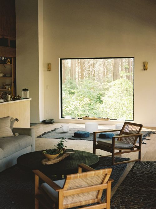 Living room interior featuring wooden chairs, a dark coffee table with greenery, and a view of trees through a large window.