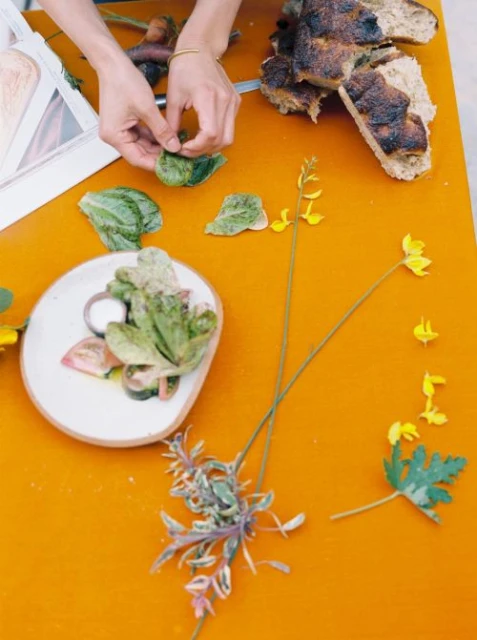 Overhead shot of food preparation: a salad being assembled with greens, bread, and flowers on an orange surface with a person's hands.