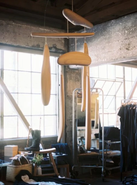 Abstract mobile sculpture hangs in a room with natural light, concrete walls, and a rack of clothes; a stool and boots rest nearby.