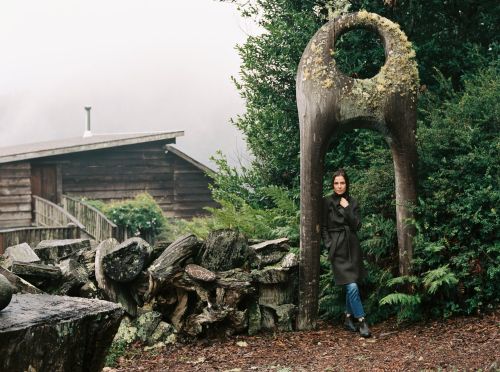 Woman in coat stands near unique wooden sculpture with foliage, by wooden cabin with foggy sky in outdoor rural setting.