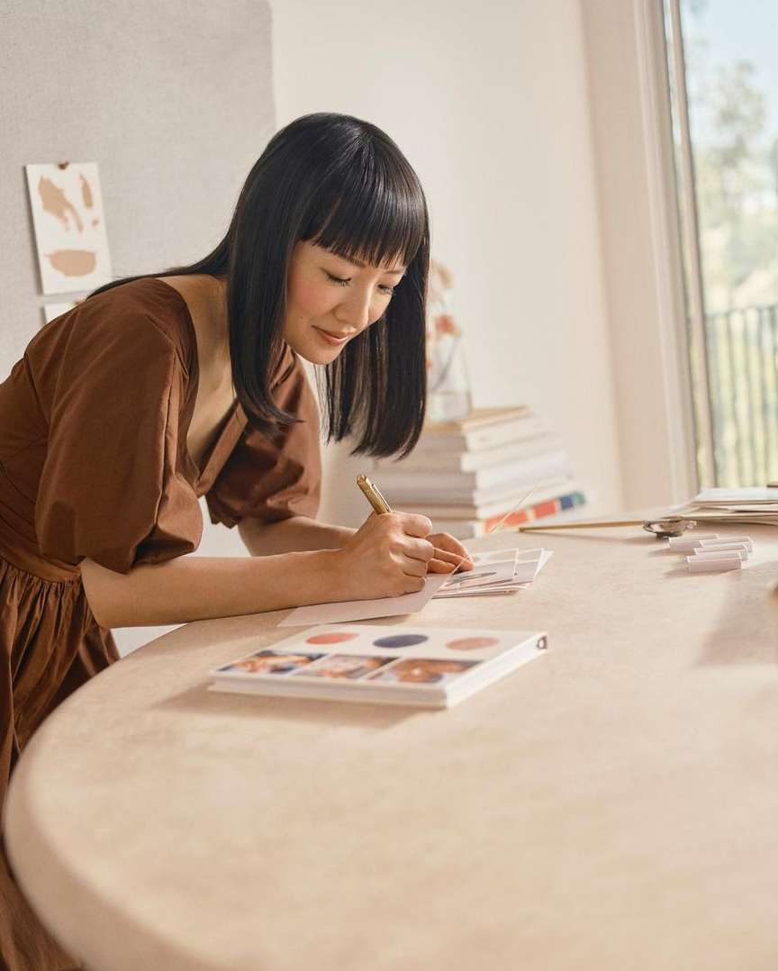 A brunette woman in a brown dress sits at a table writing in a notebook in a bright, airy room near a window and a stack of books.