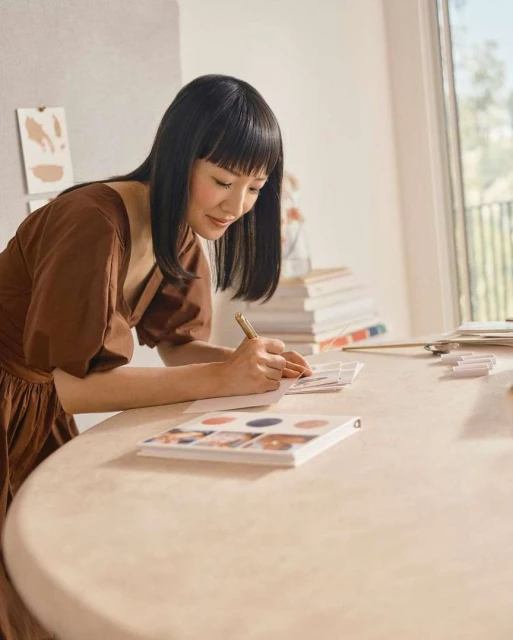 A brunette woman in a brown dress sits at a table writing in a notebook in a bright, airy room near a window and a stack of books.
