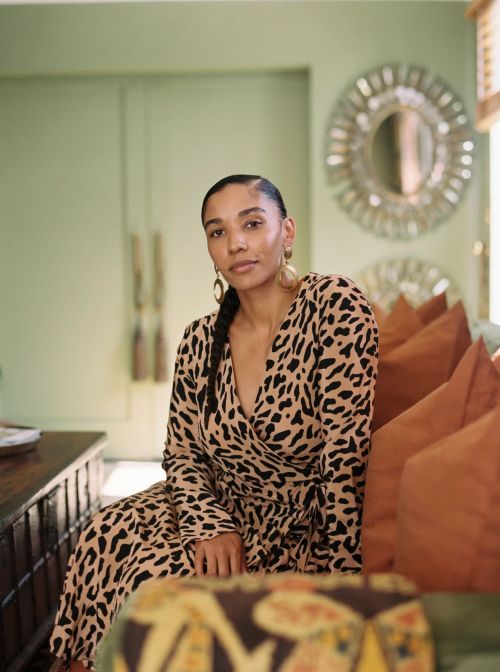 A woman in a leopard-print dress sits indoors, looking at the camera with a neutral expression, with decorative mirrors in the background.