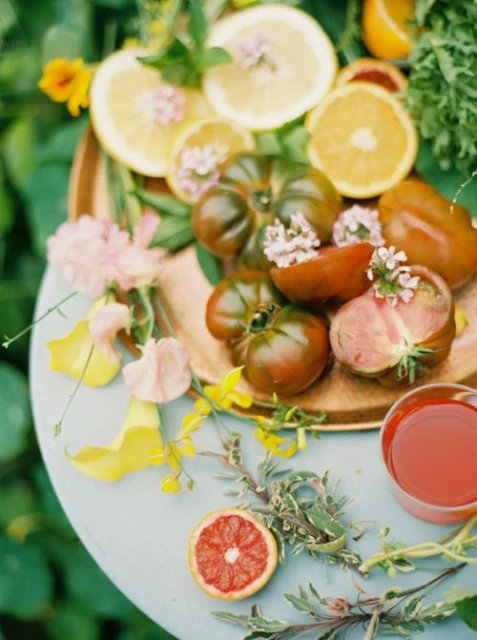 Still life of a copper tray with sliced citrus fruits, heirloom tomatoes, flowers, and greenery on a blue table against green foliage.