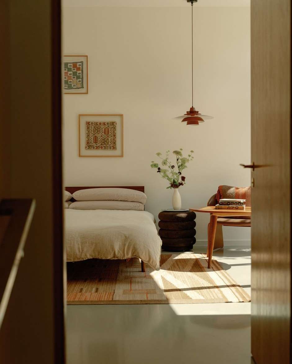 Bedroom interior with a bed, side table with flowers, and a wooden table with objects, captured through a doorway. Warm, natural light.