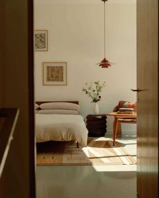 Bedroom interior with a bed, side table with flowers, and a wooden table with objects, captured through a doorway. Warm, natural light.