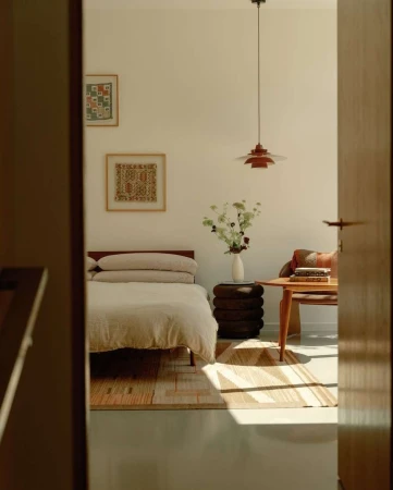 Bedroom interior with a bed, side table with flowers, and a wooden table with objects, captured through a doorway. Warm, natural light.