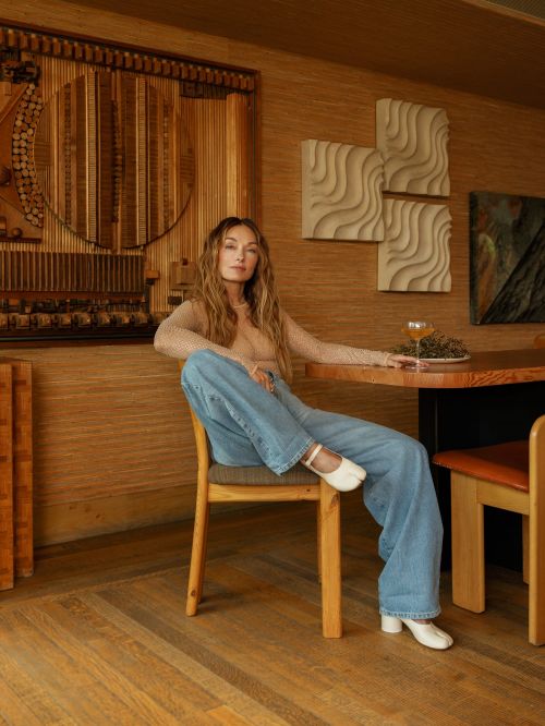 A woman with long hair poses in a chair next to a wooden table. She is wearing blue jeans and white shoes in an interior setting.