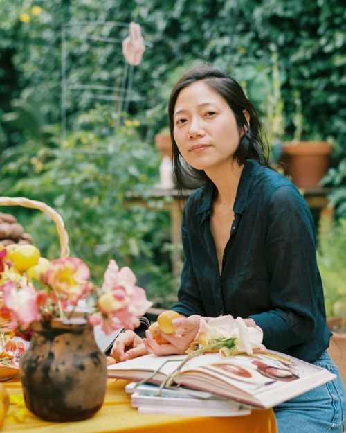 Adult woman with dark hair sits at a table in a garden, surrounded by flowers and books in a medium shot at eye-level angle.