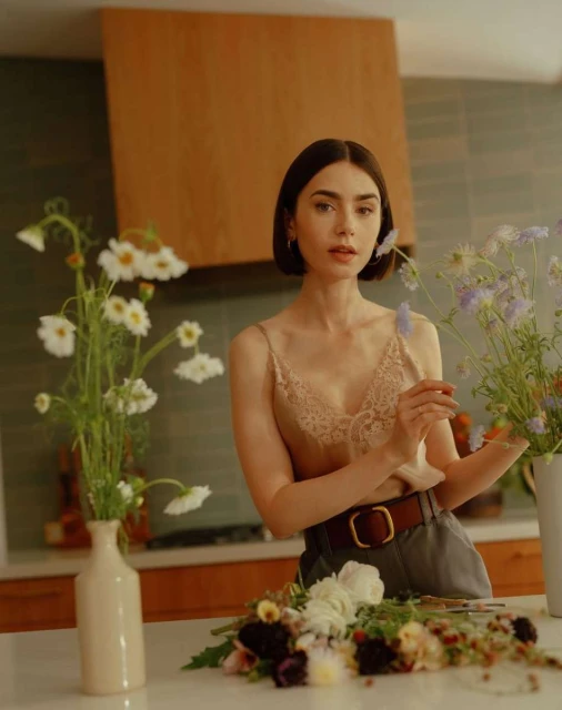 A woman arranging a flower bouquet in a kitchen, wearing a lacy top and belt, next to vases with flowers on a bright countertop.