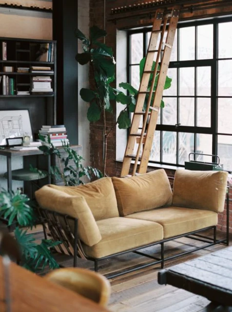 Tan-colored velvet couch with a metal frame in a loft apartment with a large window, indoor plants, a ladder, and exposed brick.