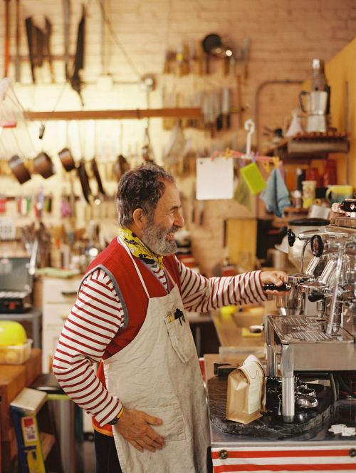 Elderly man with beard and apron operates vintage espresso machine indoors with tools hanging on the wall in background.