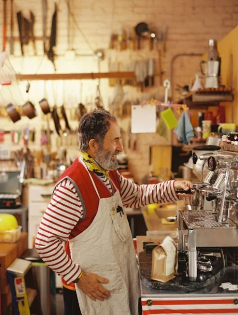 Elderly man with beard and apron operates vintage espresso machine indoors with tools hanging on the wall in background.