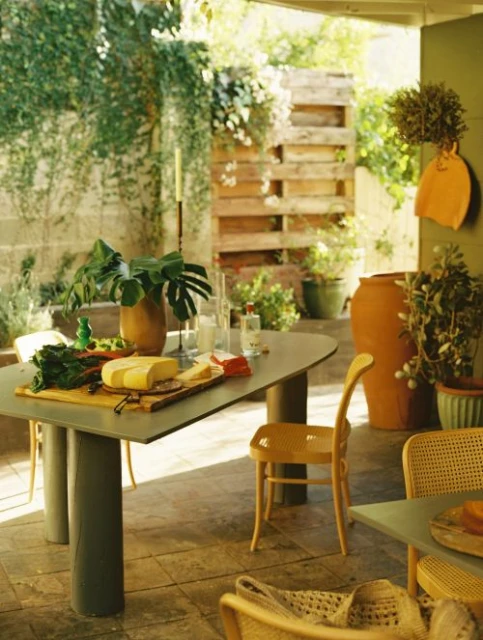 A view of a patio setting featuring a table topped with food items and surrounded by chairs, foliage, and pottery in daylight.