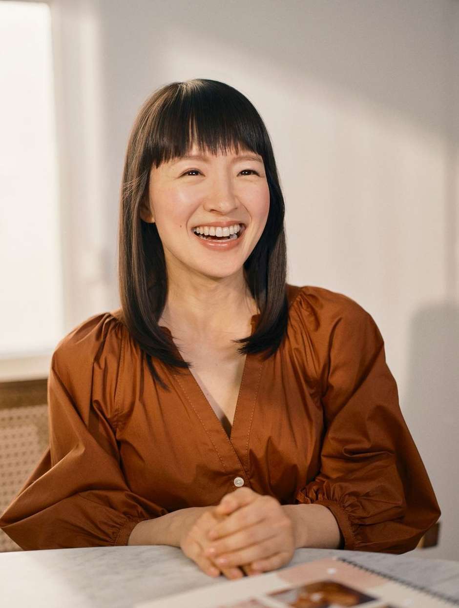 Smiling Asian woman in brown blouse with hands clasped at table in bright, neutral room with open book. Pleasant, natural, and genuine.