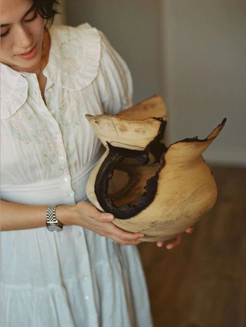 Woman in white dress holding cracked wooden vase indoors, natural light, domestic setting, art pottery, neutral color palette, close up.