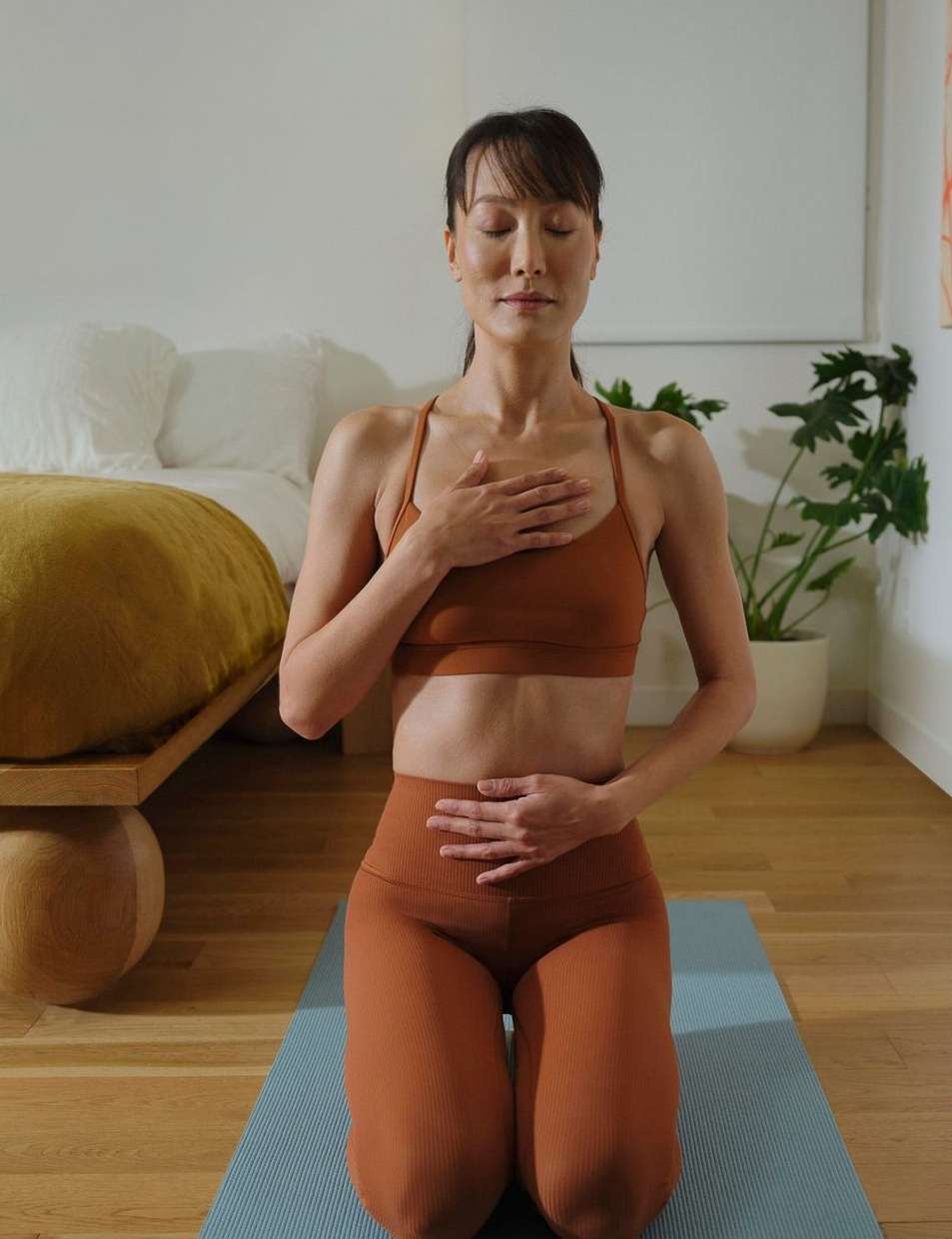 Woman practices meditation on yoga mat indoors, wearing sports bra and leggings, eyes closed, hands on chest and abdomen, calm expression.