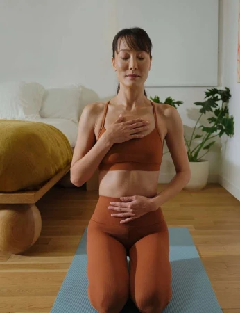 Woman practices meditation on yoga mat indoors, wearing sports bra and leggings, eyes closed, hands on chest and abdomen, calm expression.