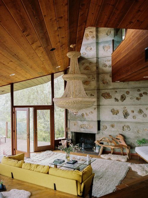 Living room interior with a yellow sofa, glass coffee table, fireplace, and large crystal chandelier, and light wood ceiling and stone wall.