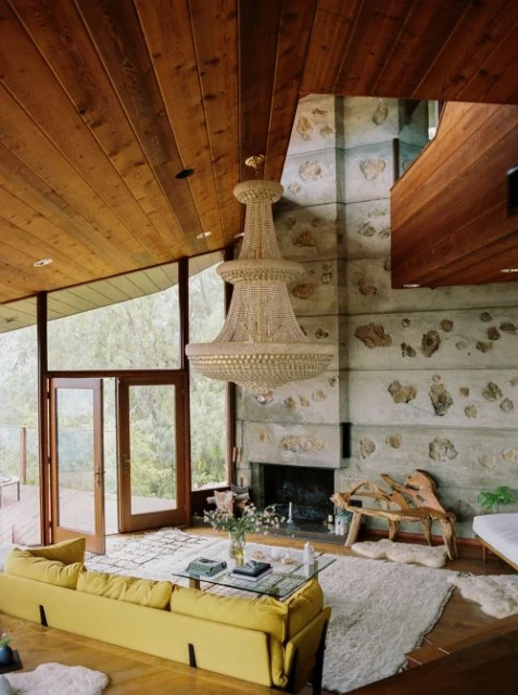 Living room interior with a yellow sofa, glass coffee table, fireplace, and large crystal chandelier, and light wood ceiling and stone wall.