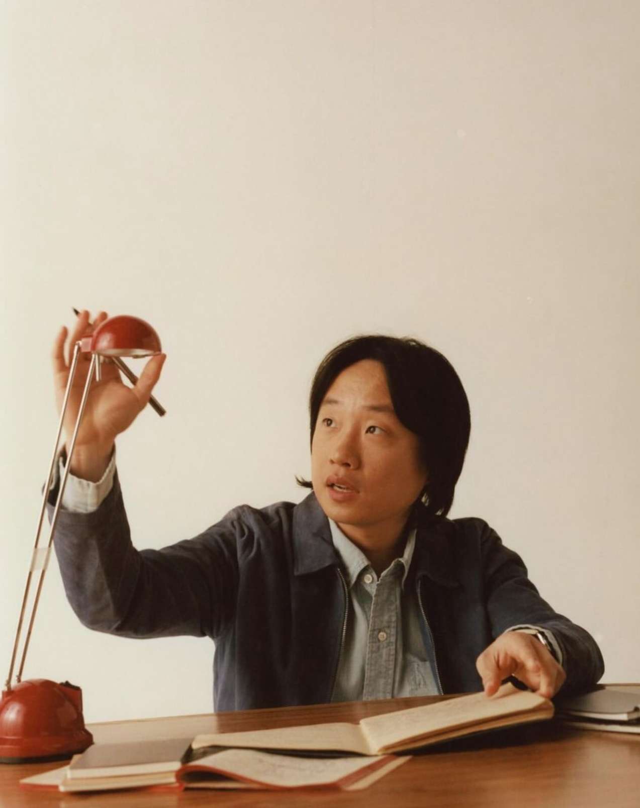 Adult male at a table with an open notebook, pen, and red adjustable lamp indoors in a medium shot with eye-level angle, soft lighting.