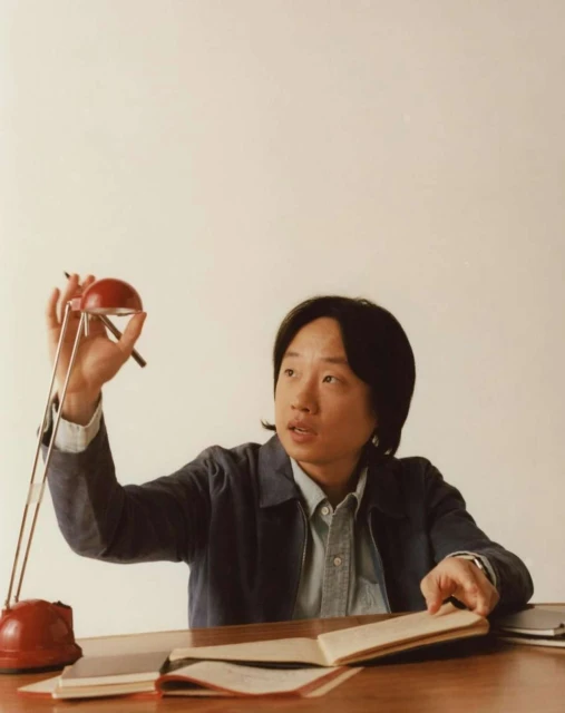 Adult male at a table with an open notebook, pen, and red adjustable lamp indoors in a medium shot with eye-level angle, soft lighting.