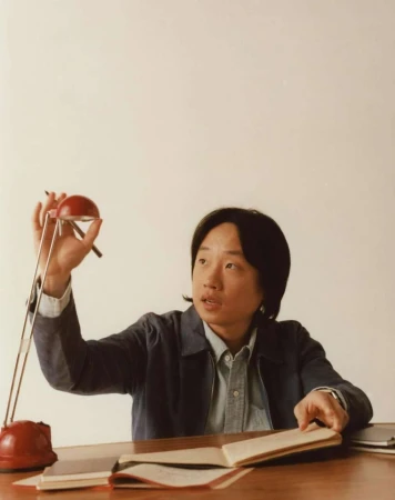 Adult male at a table with an open notebook, pen, and red adjustable lamp indoors in a medium shot with eye-level angle, soft lighting.