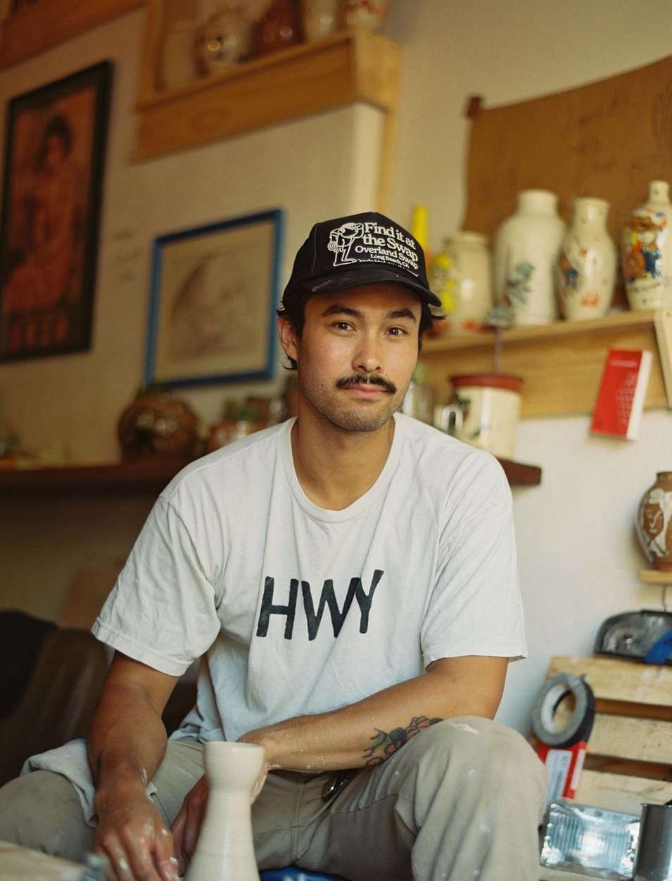 A man with a mustache and a baseball cap sits in a pottery studio. He is wearing a t-shirt and is holding a ceramic vase.