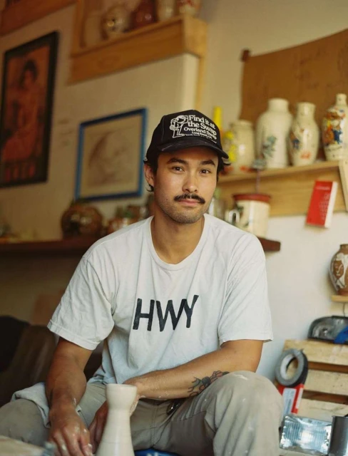 A man with a mustache and a baseball cap sits in a pottery studio. He is wearing a t-shirt and is holding a ceramic vase.